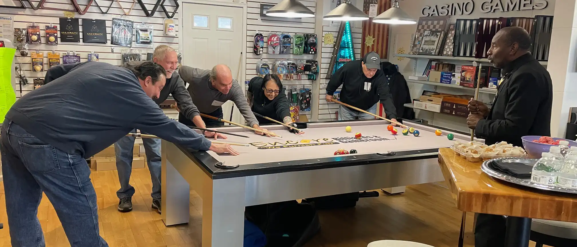 A group of diverse people enjoy playing pool together in a game room showroom featuring billiards equipment, poker sets, and various casino games.
