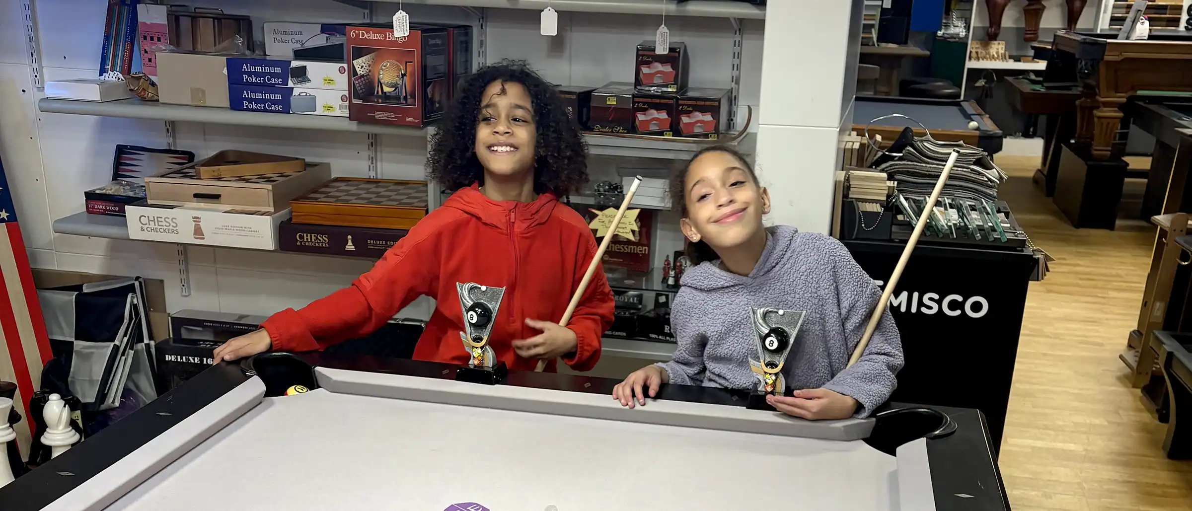 Two smiling children hold pool cues and 8-ball trophies while standing behind a white felt billiard table in a game store filled with chess sets.
