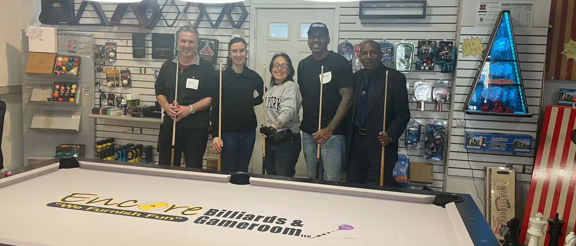 Five smiling people hold pool cues behind a white Encore Billiards & Gameroom pool table in a shop filled with billiard accessories and equipment.