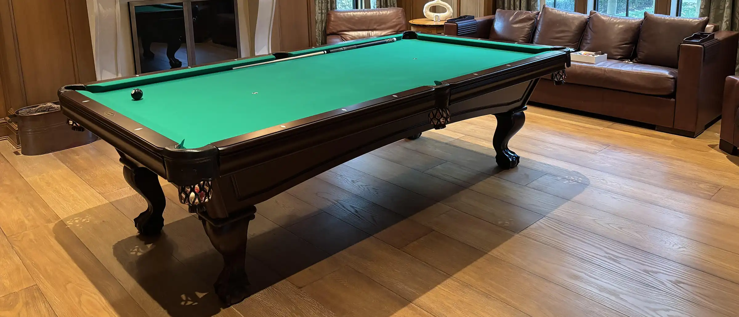 A classic dark wood billiard table with a green felt surface and ball-and-claw feet sits in a luxurious wood-paneled den with leather seating.