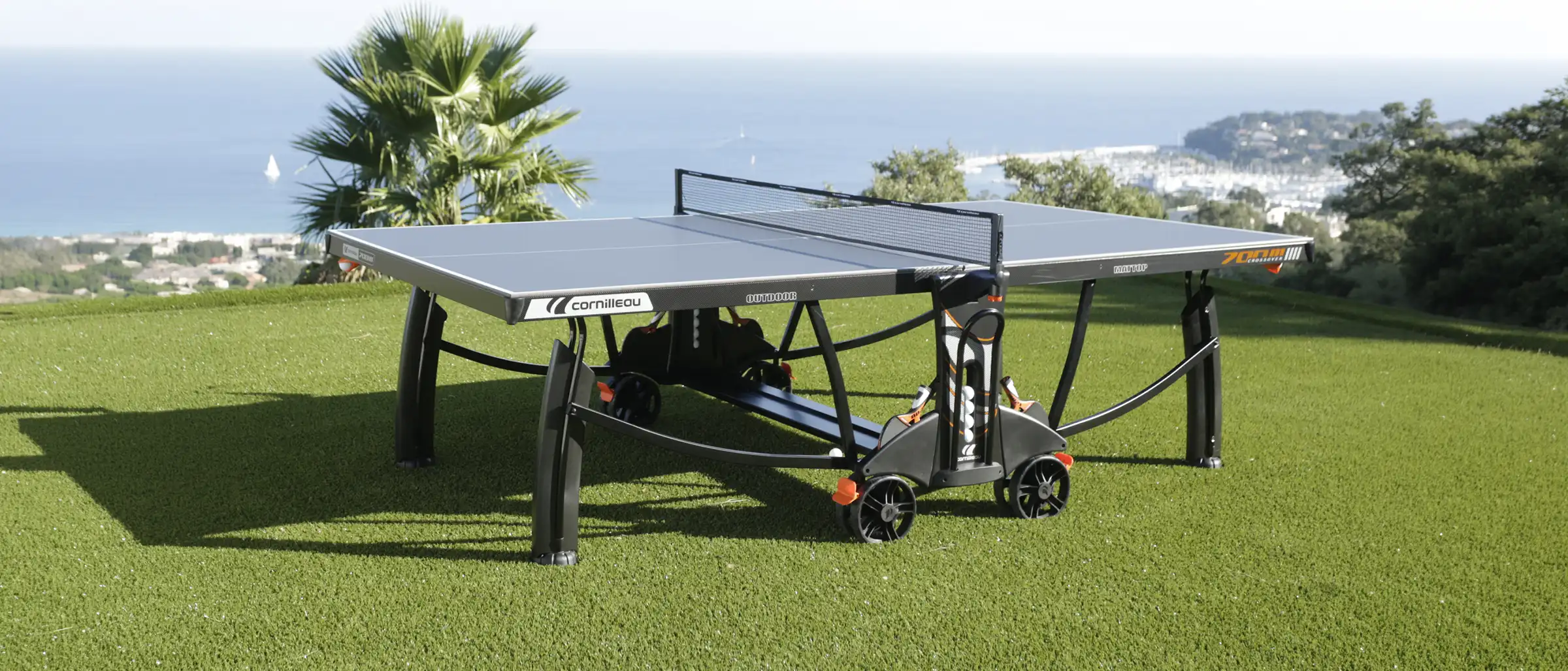 A Cornilleau outdoor table tennis table stands on a green lawn overlooking a scenic coastal view with blue ocean water and a distant harbor.
