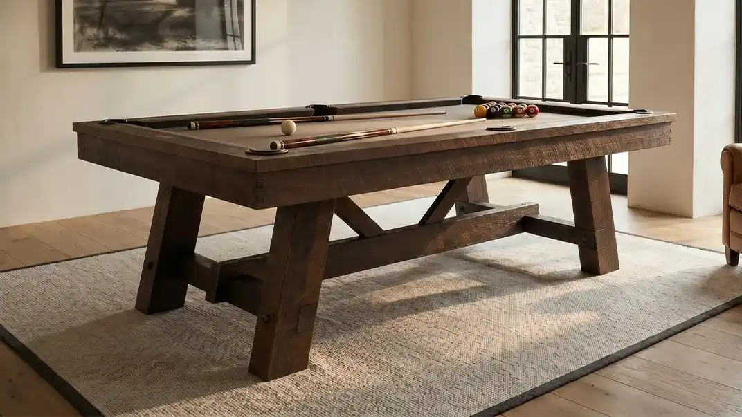 A rustic CL Bailey Tunbridge pool table made of dark weathered wood, featuring a brown felt top, billiard balls, and two pool cues.