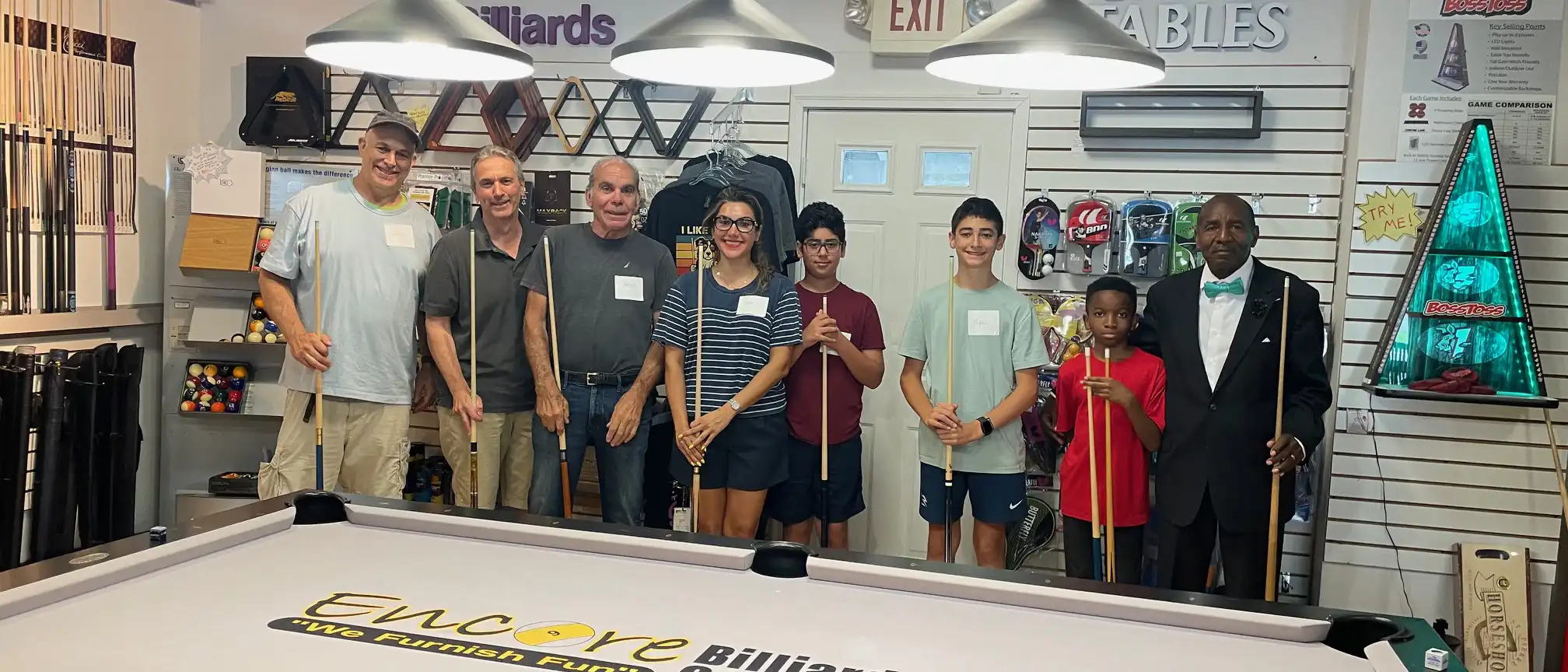 A diverse group of people standing behind an Encore Billiards pool table holding pool cues inside a billiards equipment retail store.