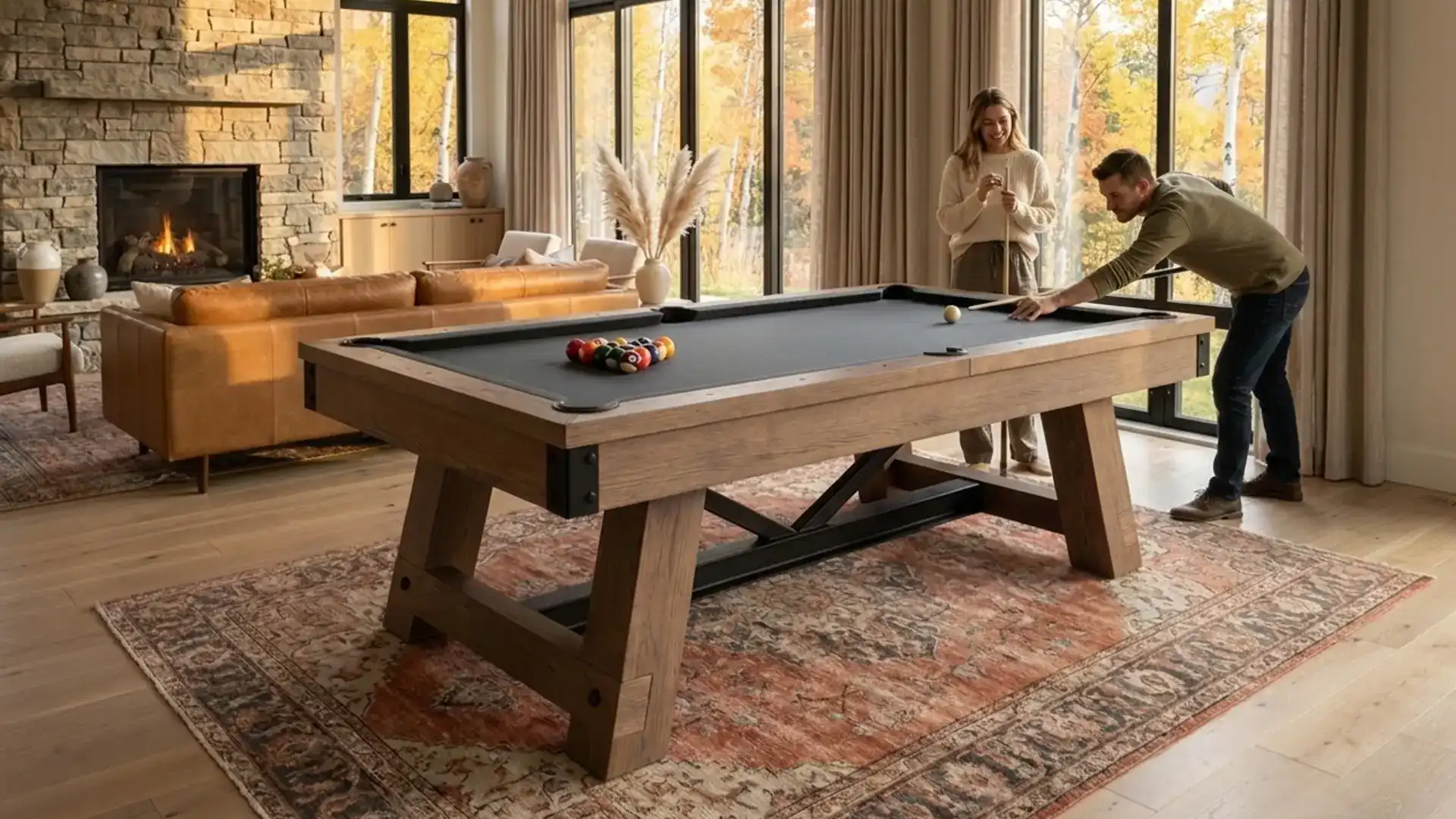 Couple playing on a rustic wooden pool table on an ornate area rug in a cozy, bright living room next to a stone fireplace.