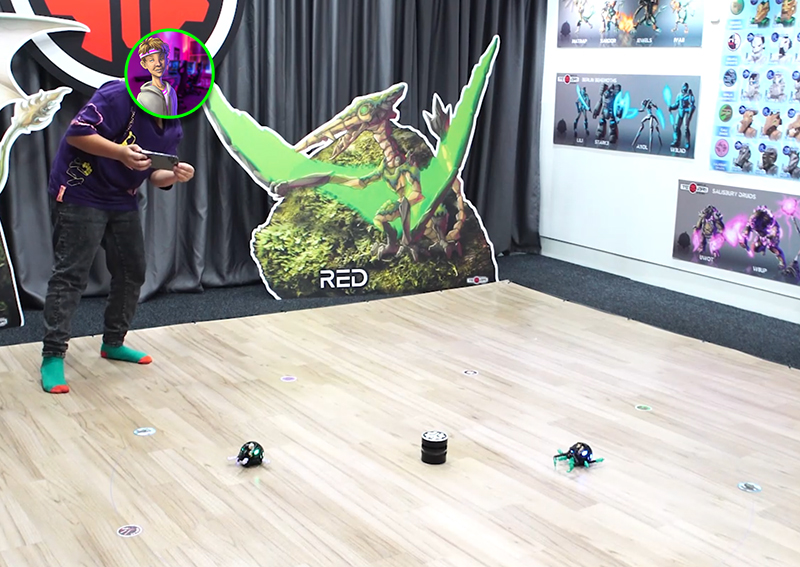 Photo of a young boy playing Temari on a mobile device with two physical mechs and a tower on the floor. His face is covered by a circular W8UP avatar.