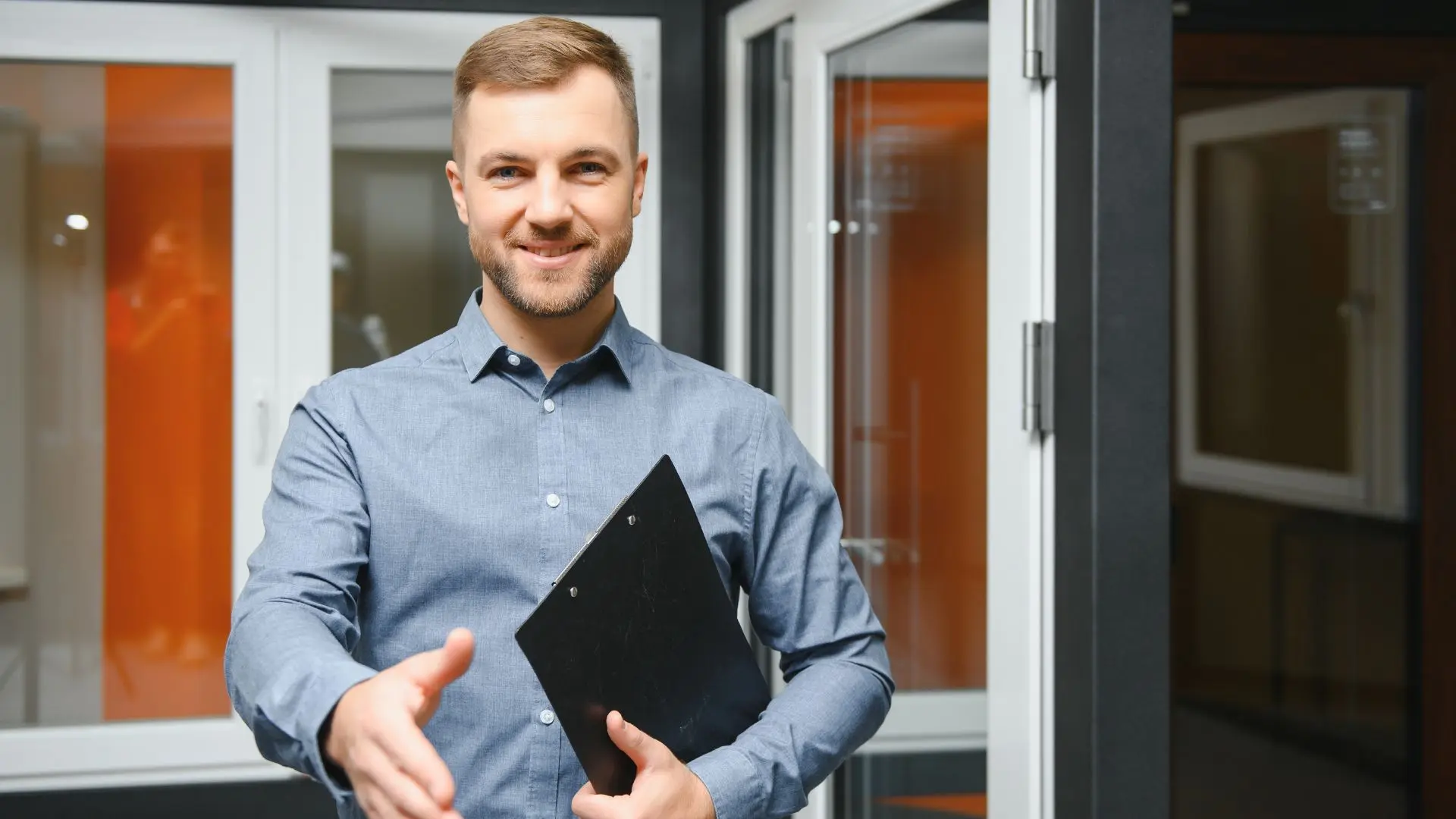 Smiling sales engineer in a blue shirt holding a clipboard and extending a hand for a handshake.