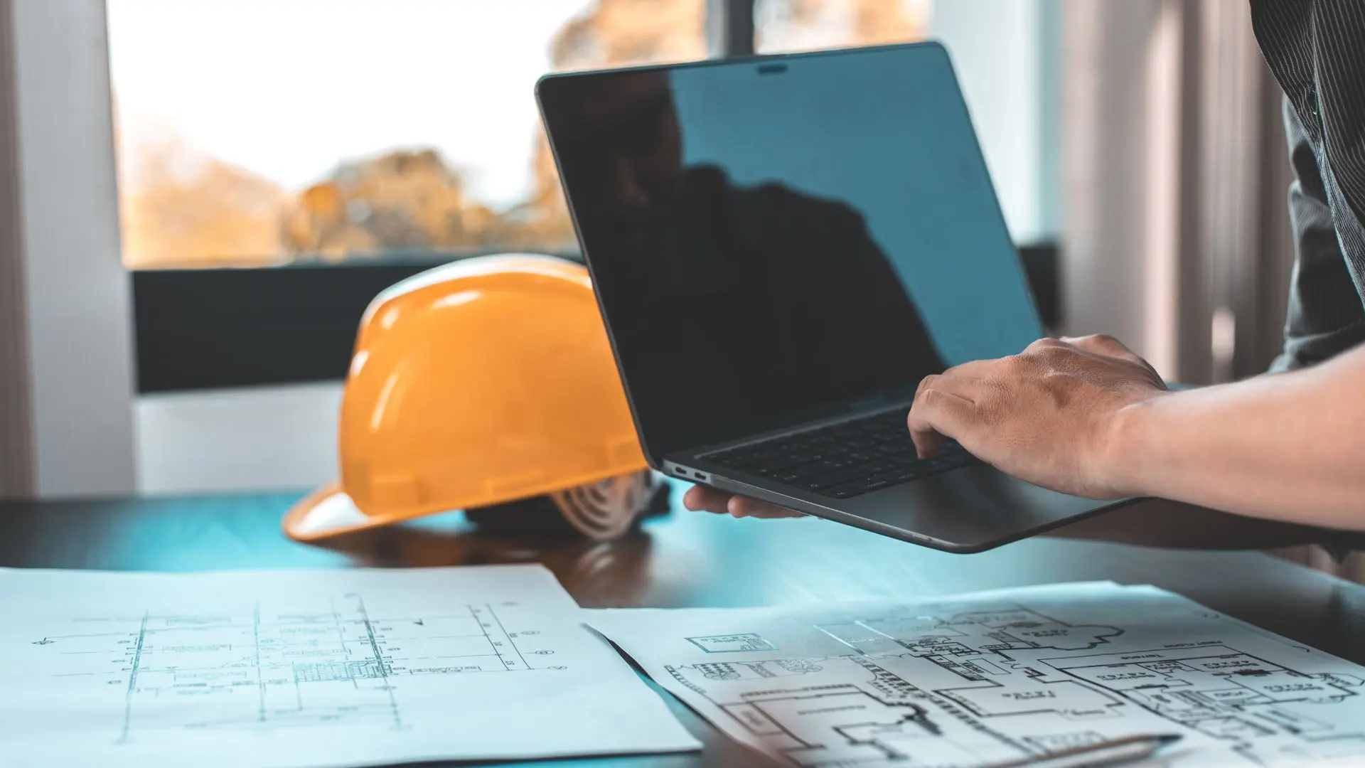 Bauingenieur:in arbeitet am Laptop an der digitalen Projektplanung mit ausgelegten Bauplänen und einem Schutzhelm auf dem Schreibtisch zur Bewältigung des Ingenieurmangels in der Industrie.