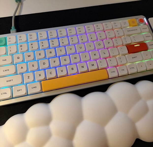 Customer-uploaded image. The image represents: A white mechanical keyboard with colorful RGB backlighting and white keycaps, photographed from above with a blurred hand visible in the foreground