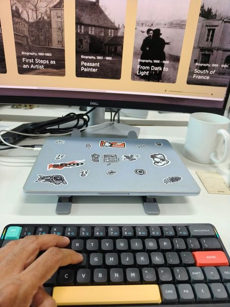 Customer-uploaded image. The image represents: A laptop covered in various stickers sits on a desk stand in front of a Dell monitor displaying biographical timeline images, with a hand positioned on a colorful keyboard and a white coffee mug nearby