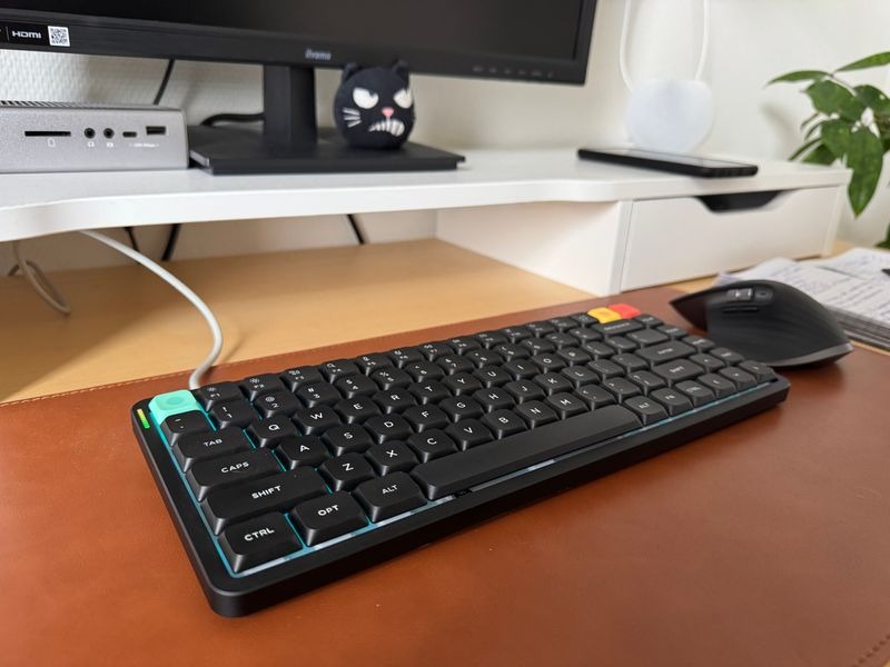 Customer-uploaded image. The image represents: A black mechanical keyboard with blue accent lighting sits on a wooden desk next to a wireless mouse, with a computer monitor and decorative items visible in the background