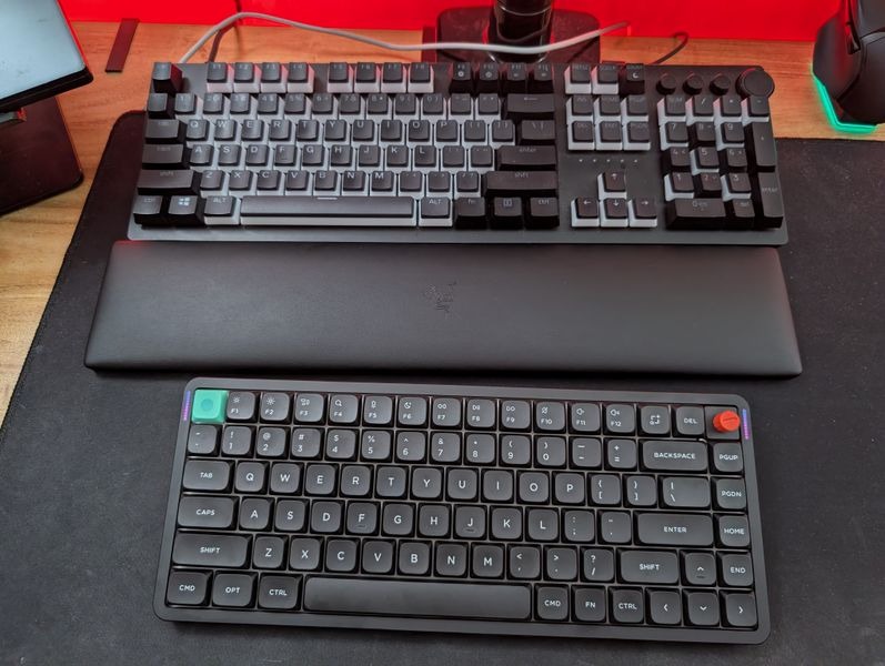 Customer-uploaded image. The image represents: A desk setup showing three keyboards - a large mechanical keyboard at the top, a laptop computer in the middle, and a compact wireless keyboard at the bottom, all arranged on a dark mouse pad on a wooden desk surface.