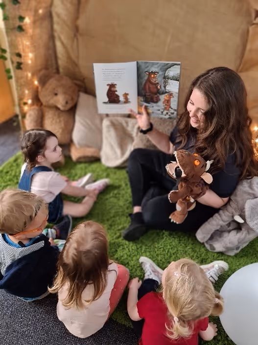 A woman reads a storybook to four young children sitting on a green carpet, with plush toys nearby.