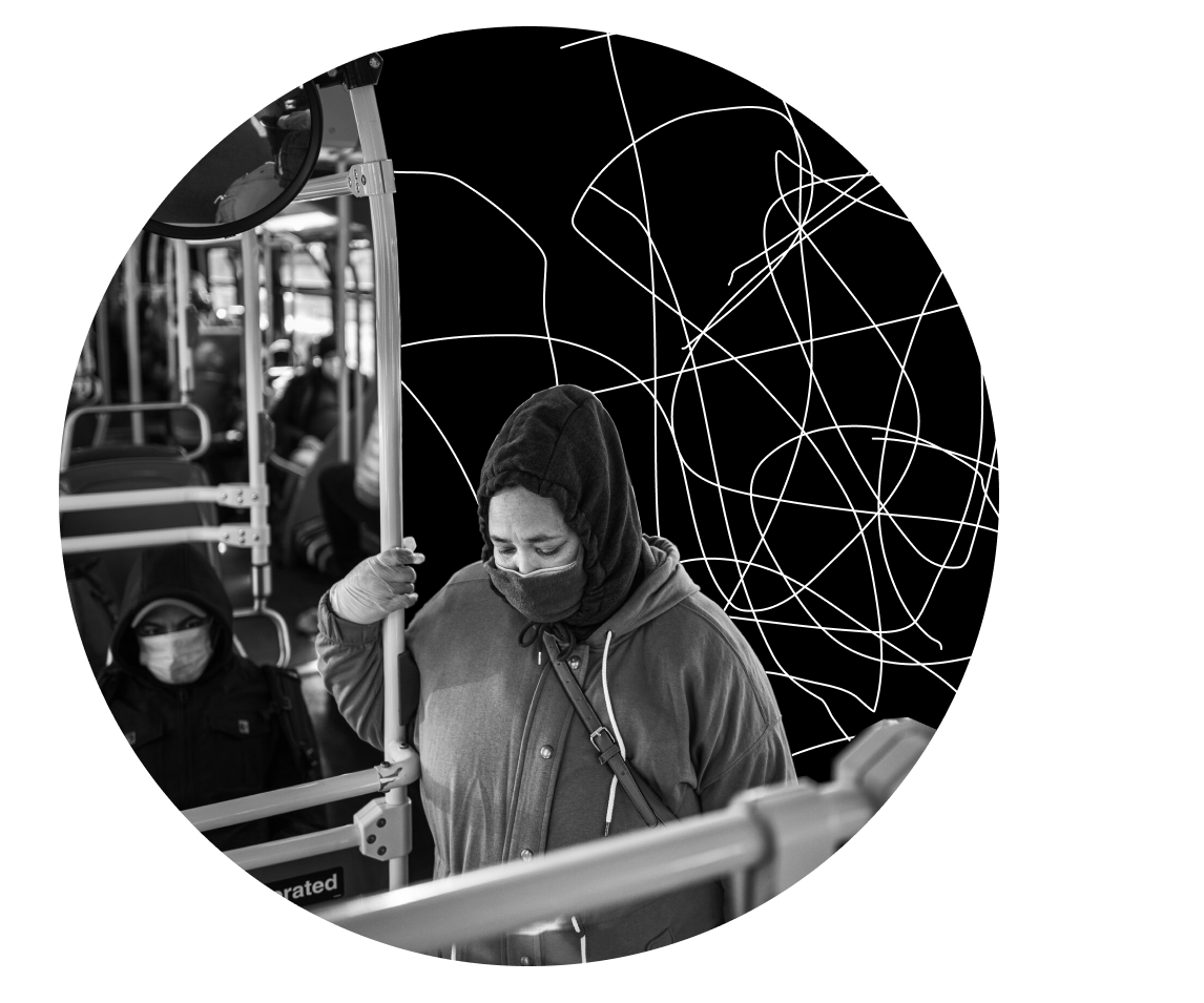 Person wearing a hood and face mask holding a pole inside a bus, with another masked passenger seated in the background.