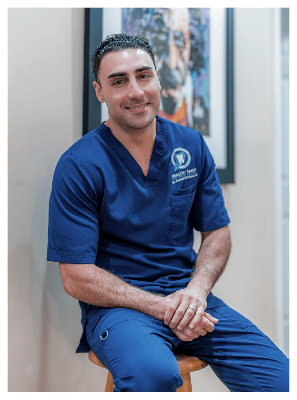 Dr. Joseph Cipriano, DDS, Staten Island dentist, smiling on a wooden stool in his Healthy Smiles scrubs