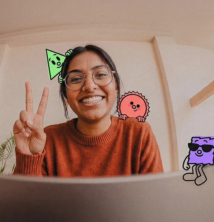 Smiling woman wearing glasses and a rust-colored sweater shows a peace sign with cartoon shapes around her head.