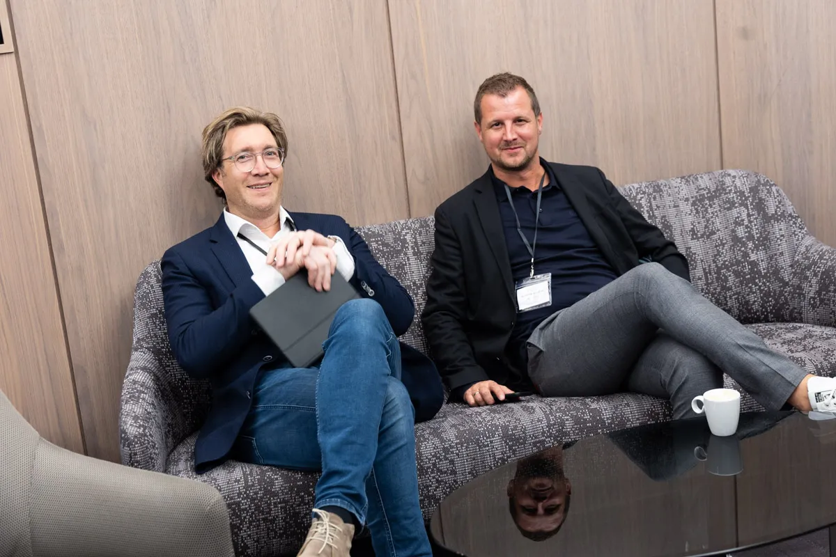 2 Männer im Business Casual Look sitzen auf grauer Couch und unterhalten sich