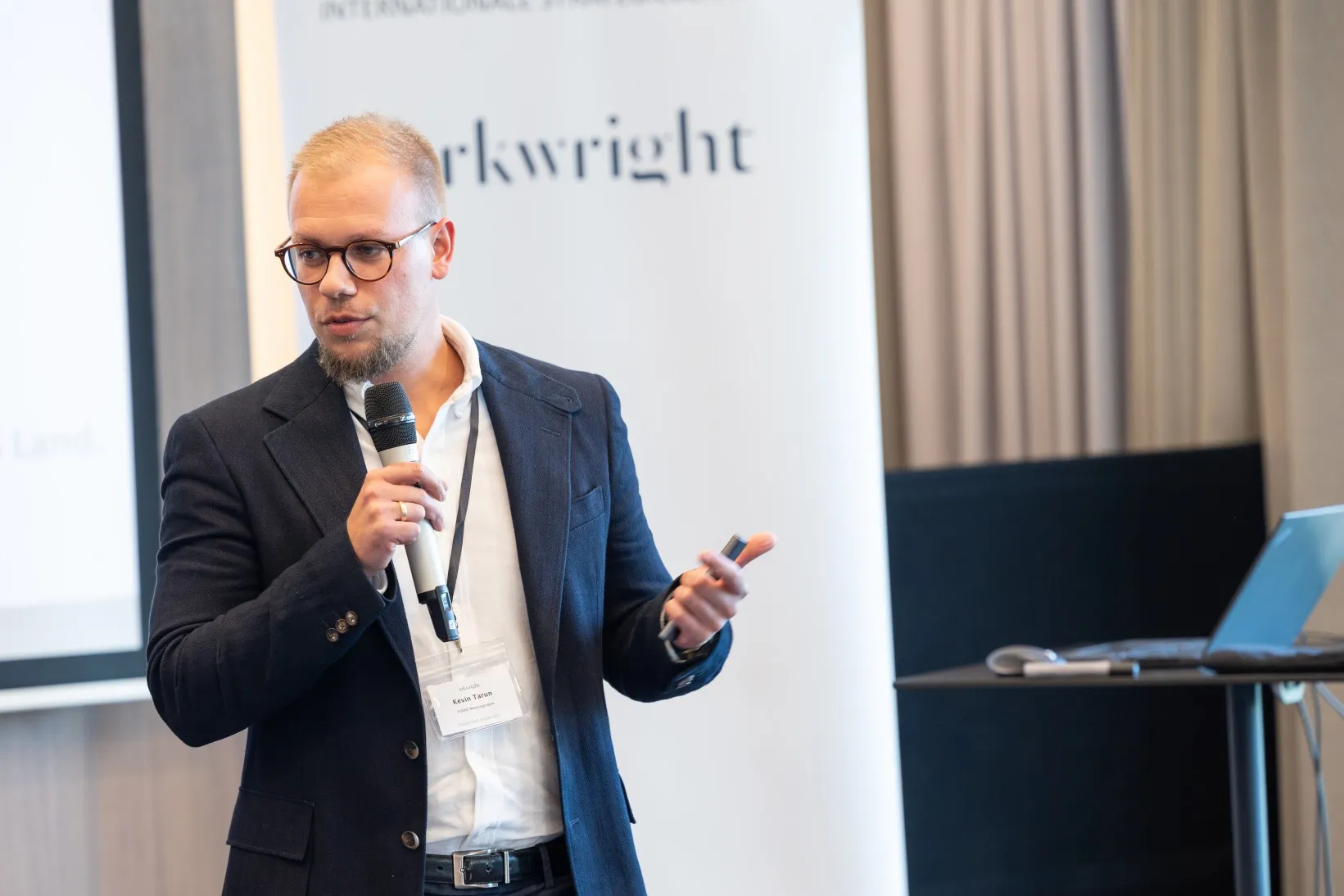 Junger Mann in blauem Sakko steht in einem Konferenzraum vorn und hält einen Vortrag