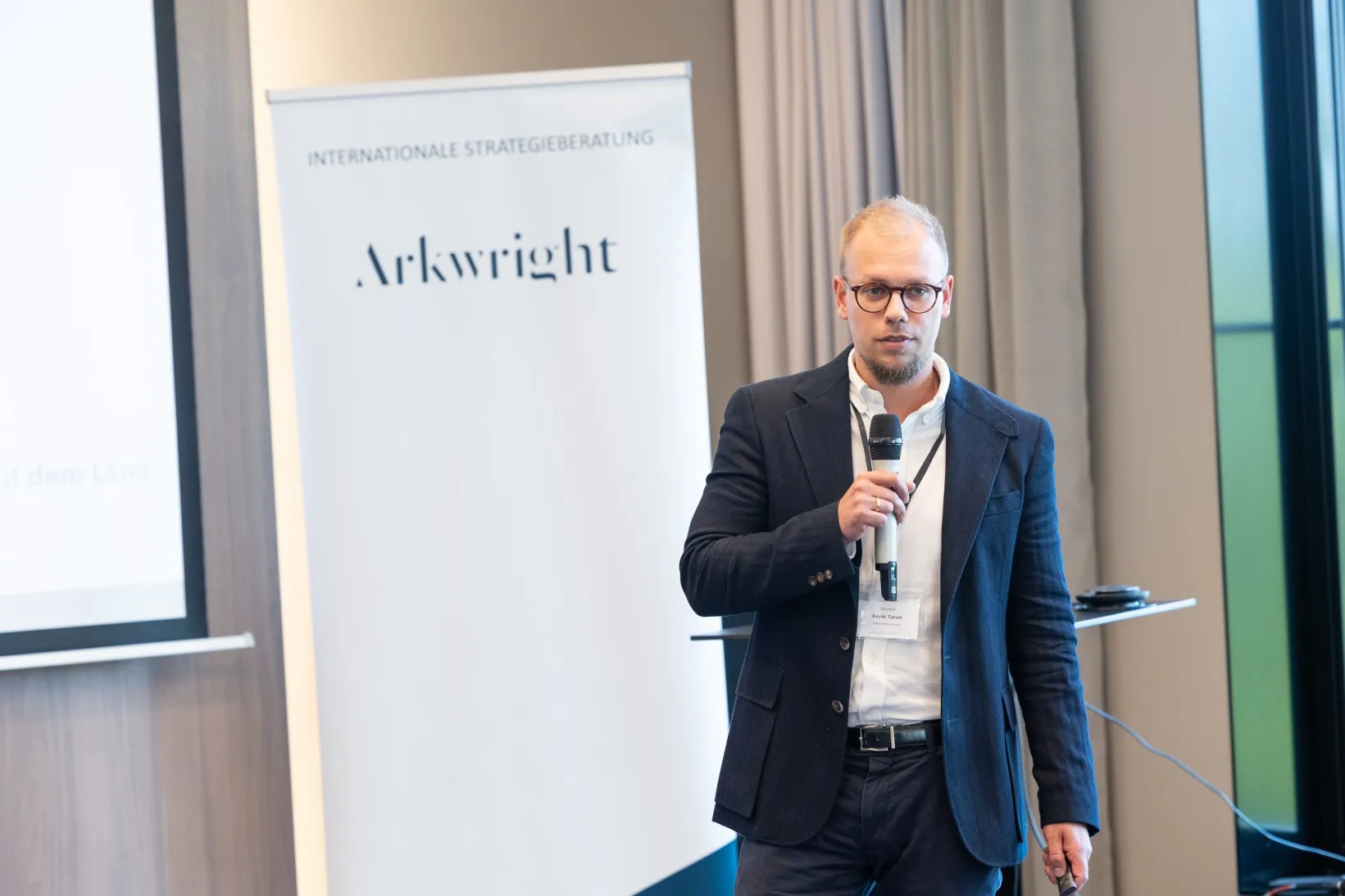 Junger Mann in blauem Sakko steht in einem Konferenzraum vorn und hält einen Vortrag