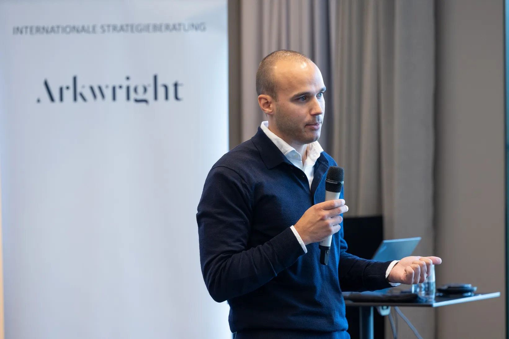 Mann in blauem Sakko steht in einem Konferenzraum vorn und hält einen Vortrag