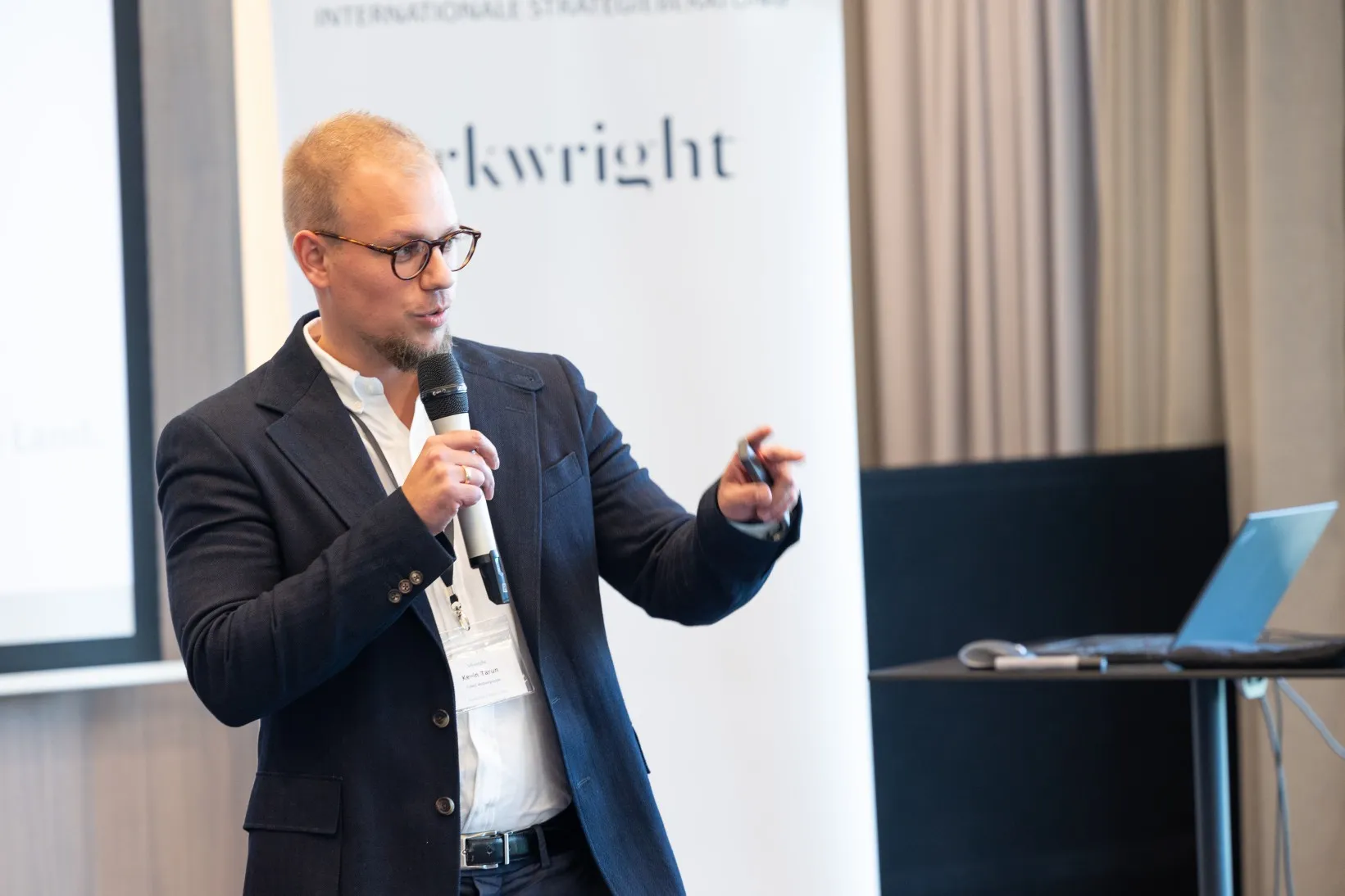 Junger Mann in blauem Sakko steht in einem Konferenzraum vorn und hält einen Vortrag