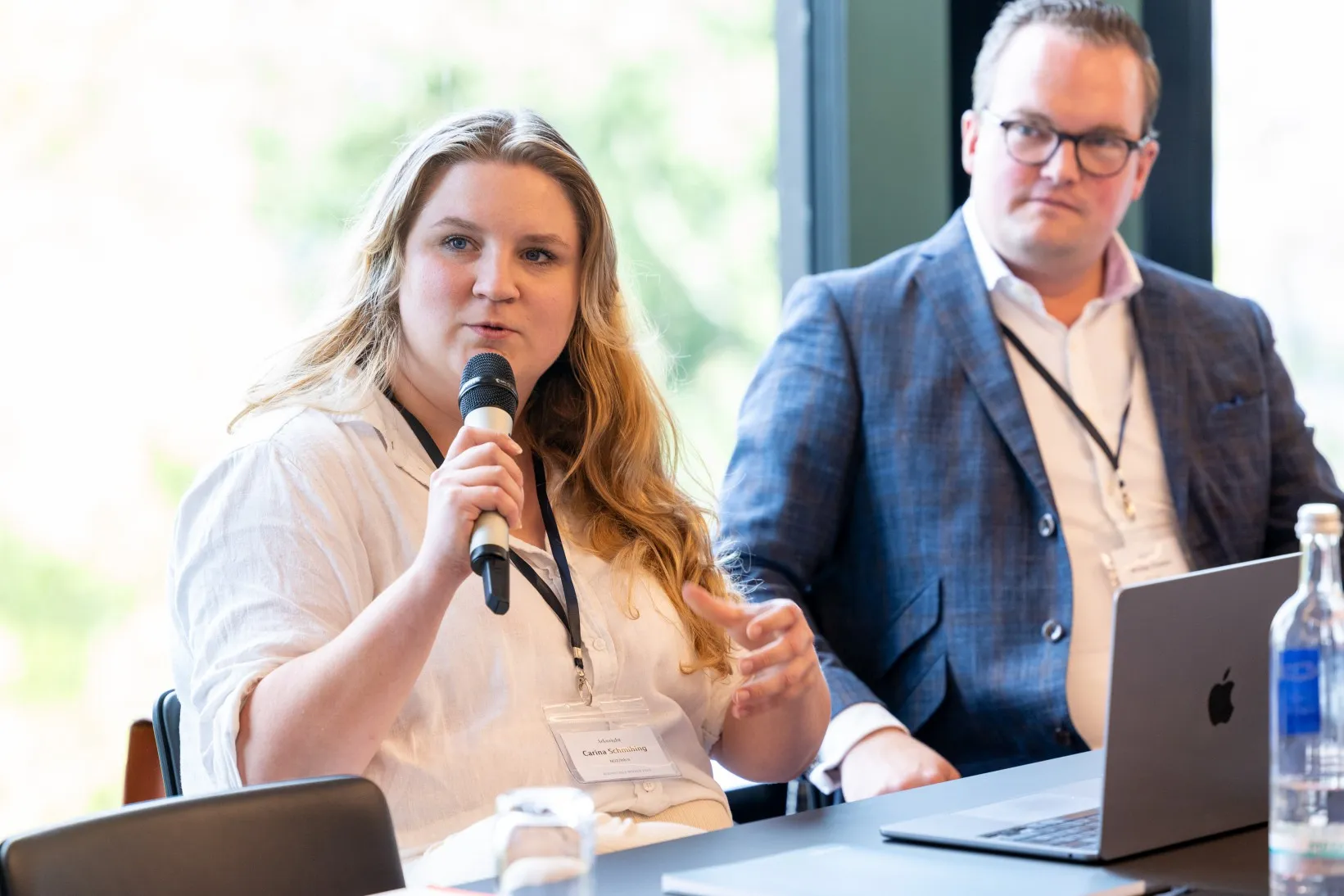 Frau in weißem Hemd hält Mikro in der Hand und sitzt an einem Konferenztisch