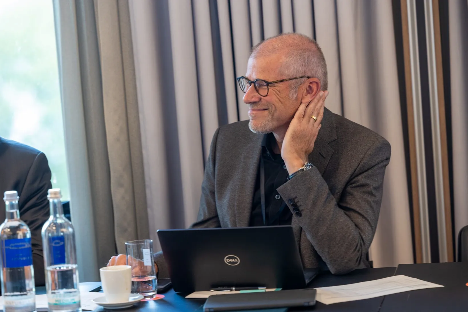 Mann in grauem Sakko und Brille sitzt lachend an seinem Laptop