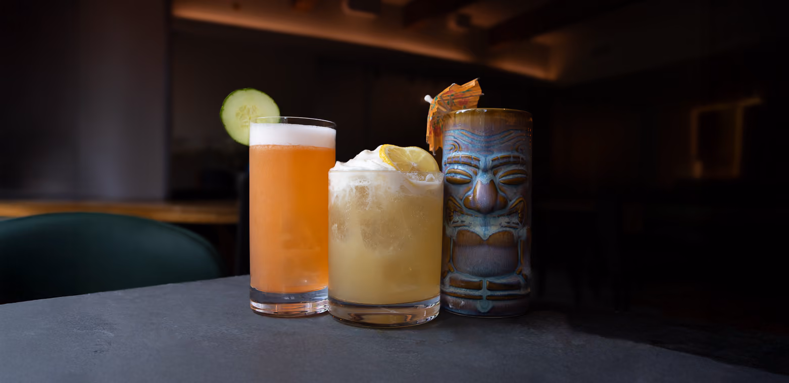 Three cocktails on dark slate: an orange drink with a cucumber slice, a pale drink with lemon, and a tiki mug