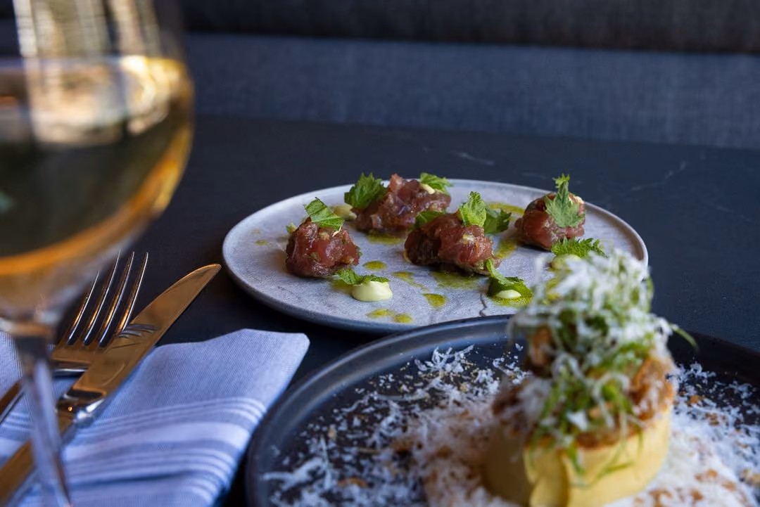 Plate of small dark red mounds with herbs and green sauce, beside a dish with shaved cheese and a glass of white wine