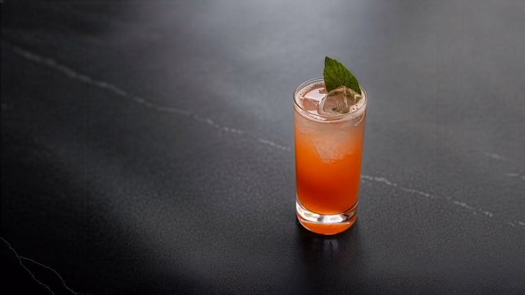 Tall orange-red cocktail with ice and a basil leaf on a dark stone counter