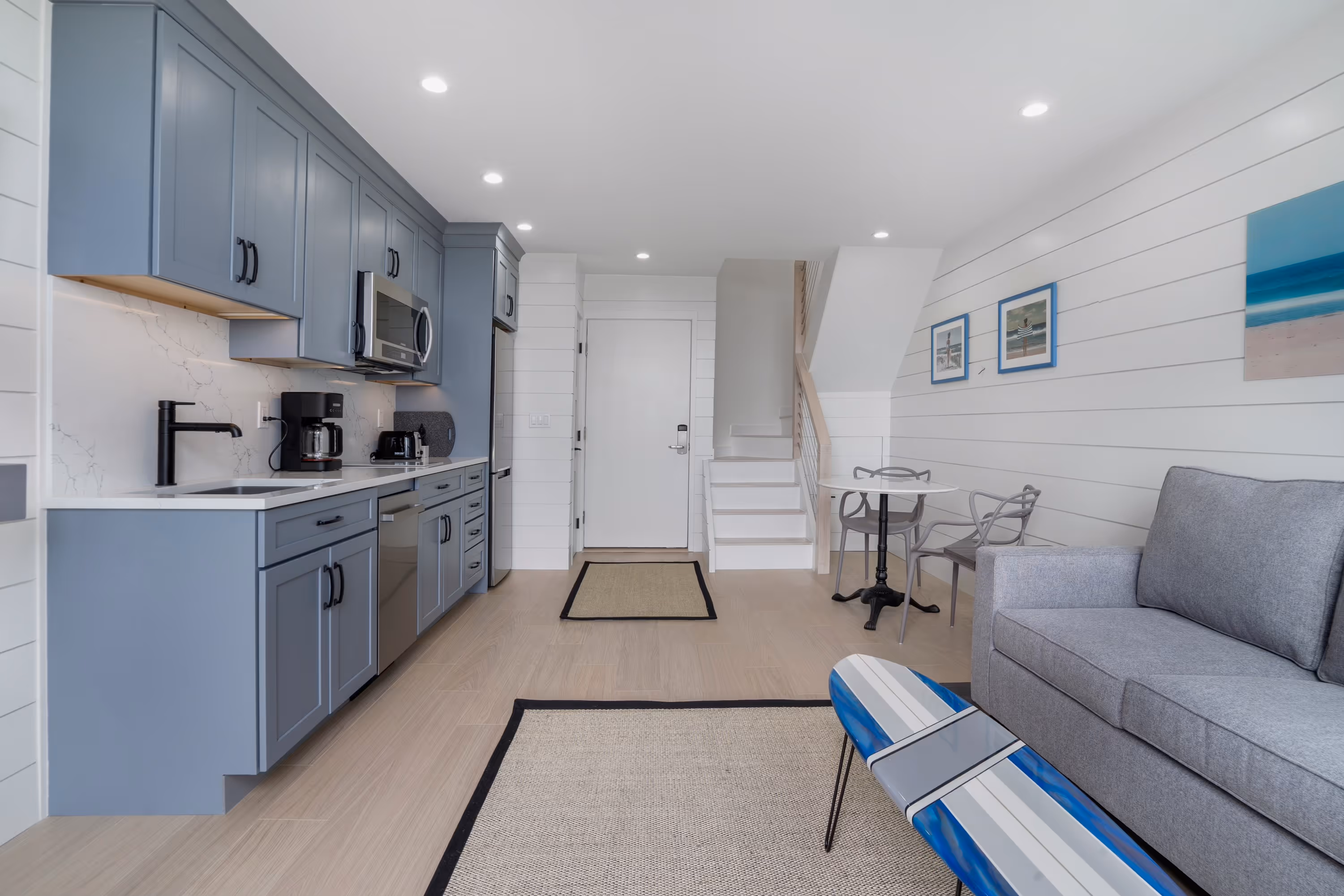 Modern open-concept kitchen and living area with blue cabinets, gray sofa, small dining table, and surfboard coffee table.