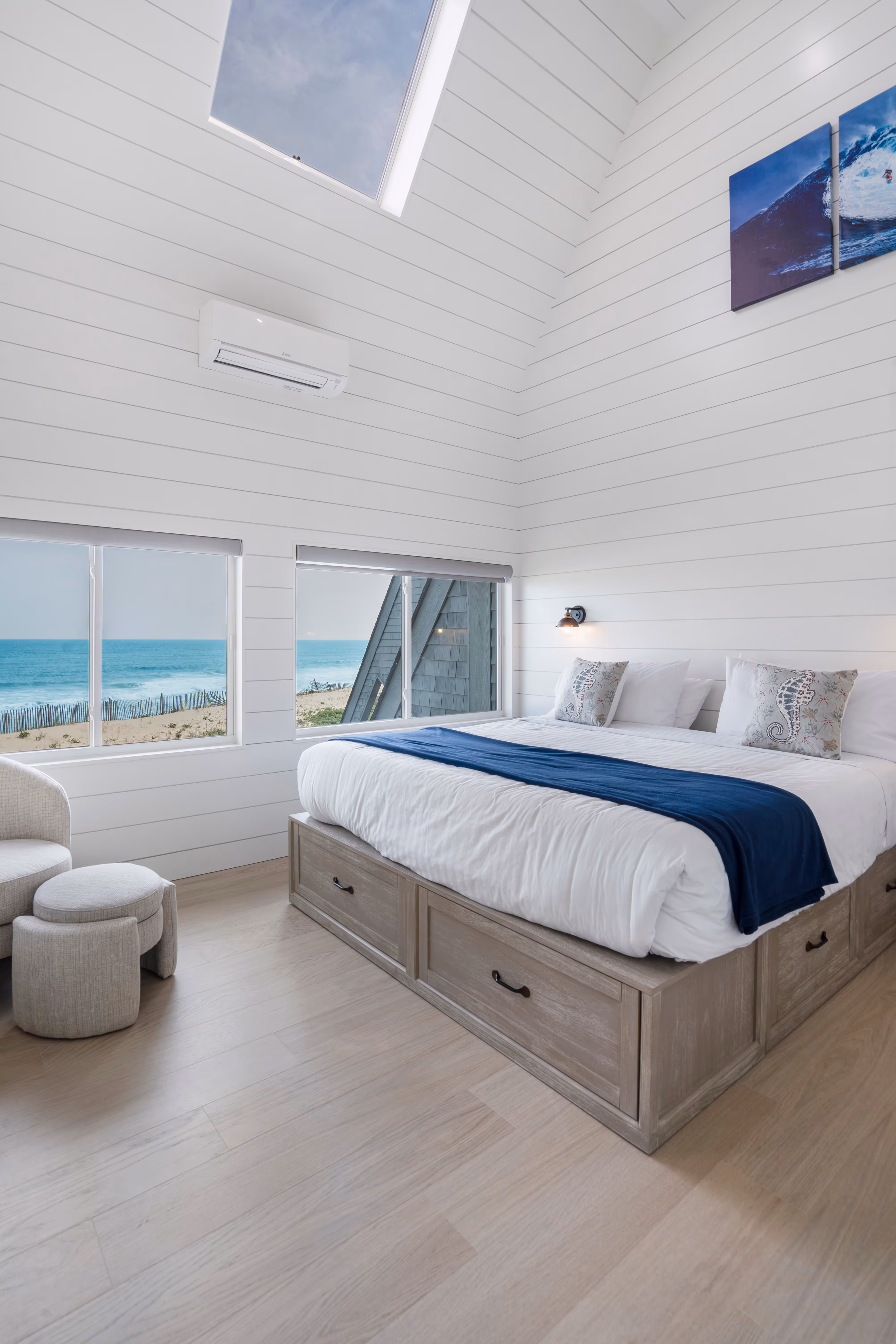 Bright bedroom with large bed, blue throw, seahorse pillows, and windows showcasing an ocean view.