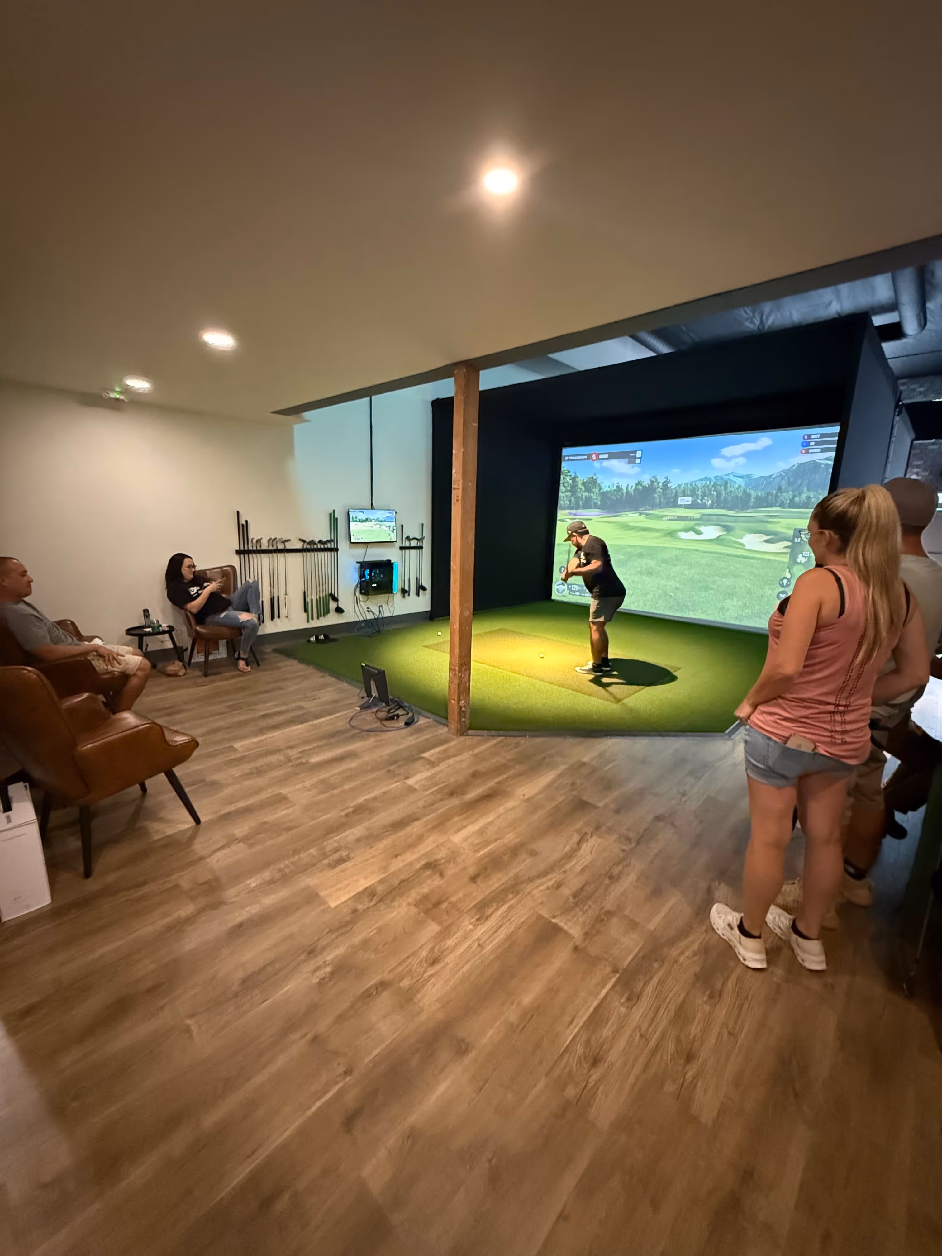 Indoor golf simulator room with a man preparing to swing a club on artificial turf while others watch seated on leather chairs and standing.