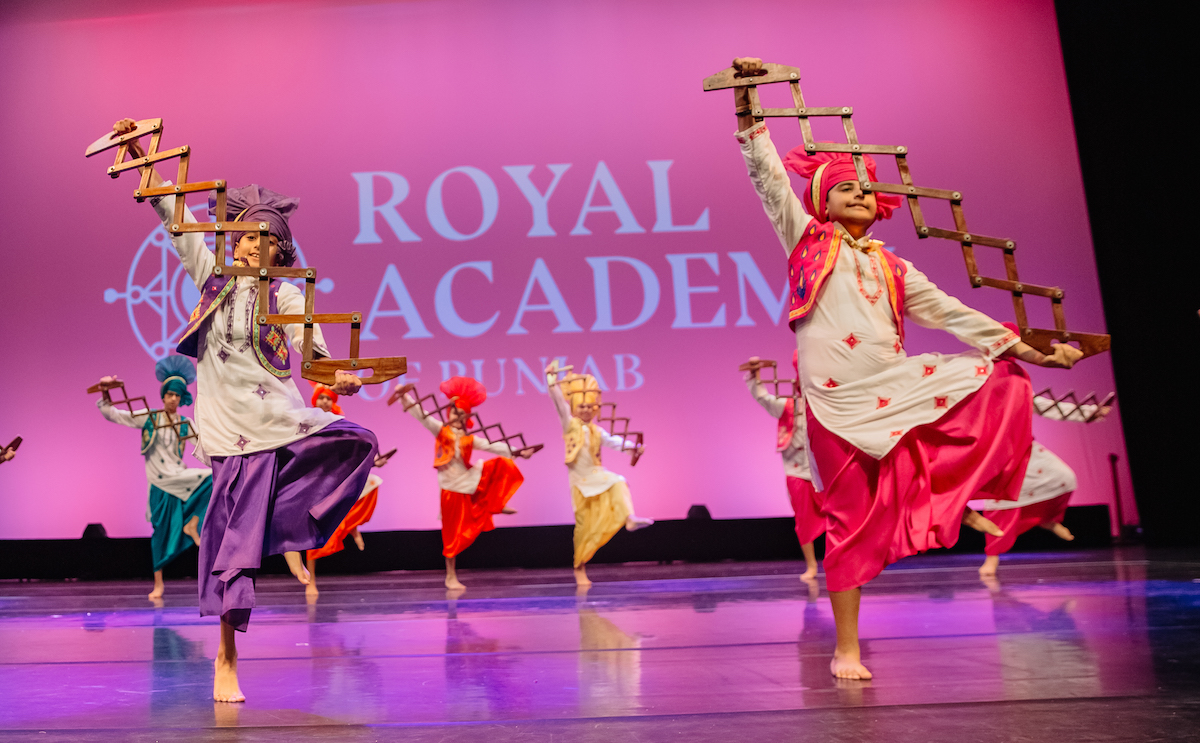 Group of children performing traditional Punjabi dance on stage wearing colorful attire and holding wooden props with a purple background and Royal Academy Punjab logo.