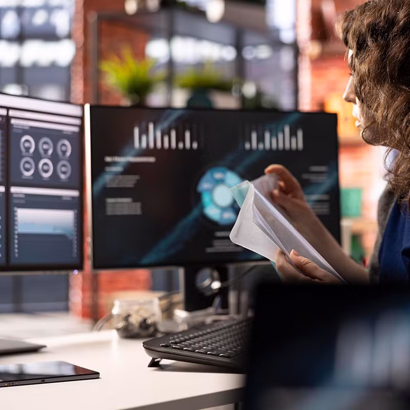 A woman reviews documents while working at a desk with dual monitors displaying dashboards, charts, and analytics, symbolizing master data management.