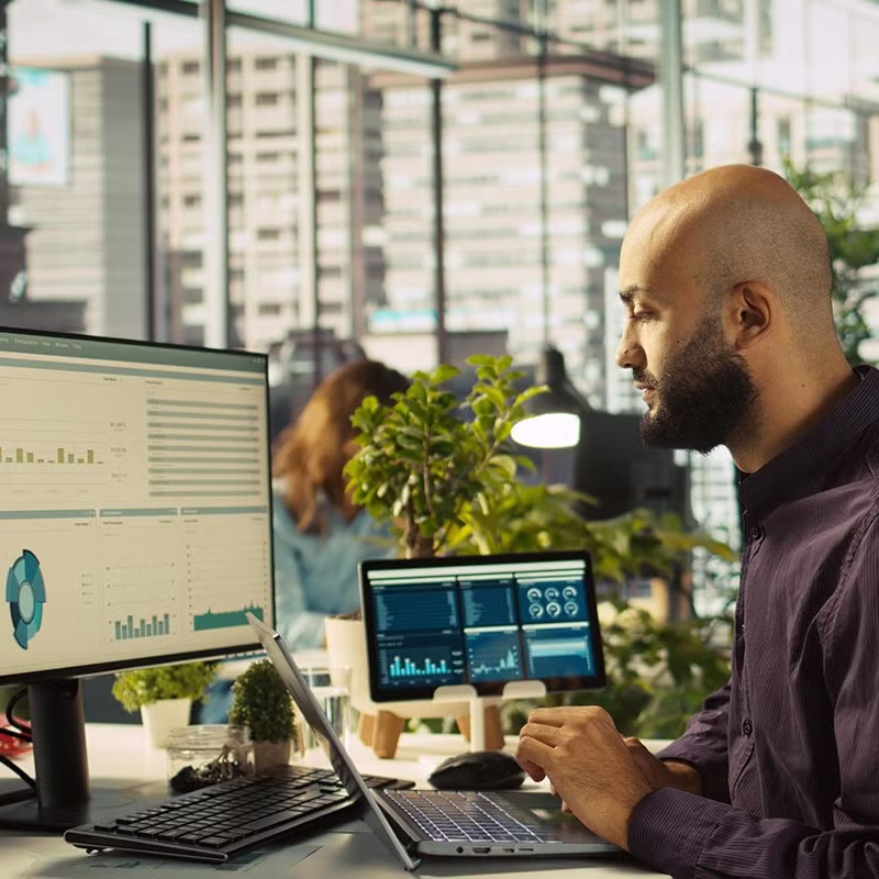 A man works at a desk with multiple screens displaying charts, graphs, and dashboards in a modern office environment.