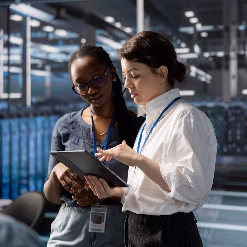 Two women stand in a modern data center reviewing information on a tablet, representing enterprise data integration and collaboration.