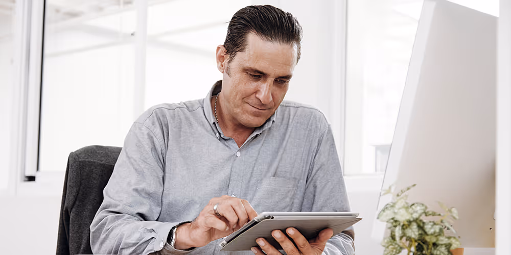 Man using a tablet at his desk in a modern office, focused on reviewing or entering information beside a desktop computer.