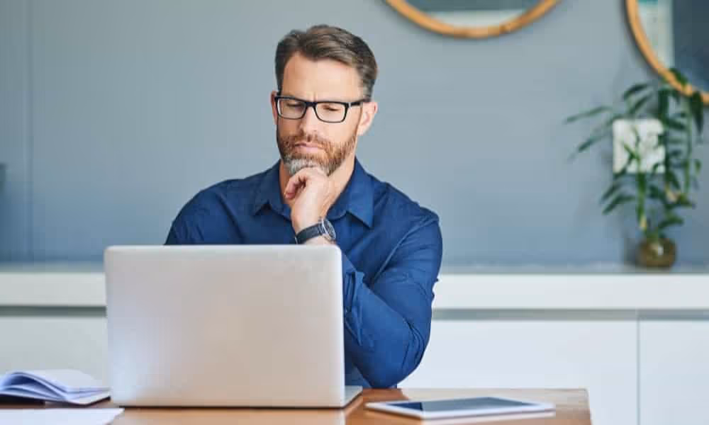 Man with glasses and beard thoughtfully looking at his laptop screen in a modern office.