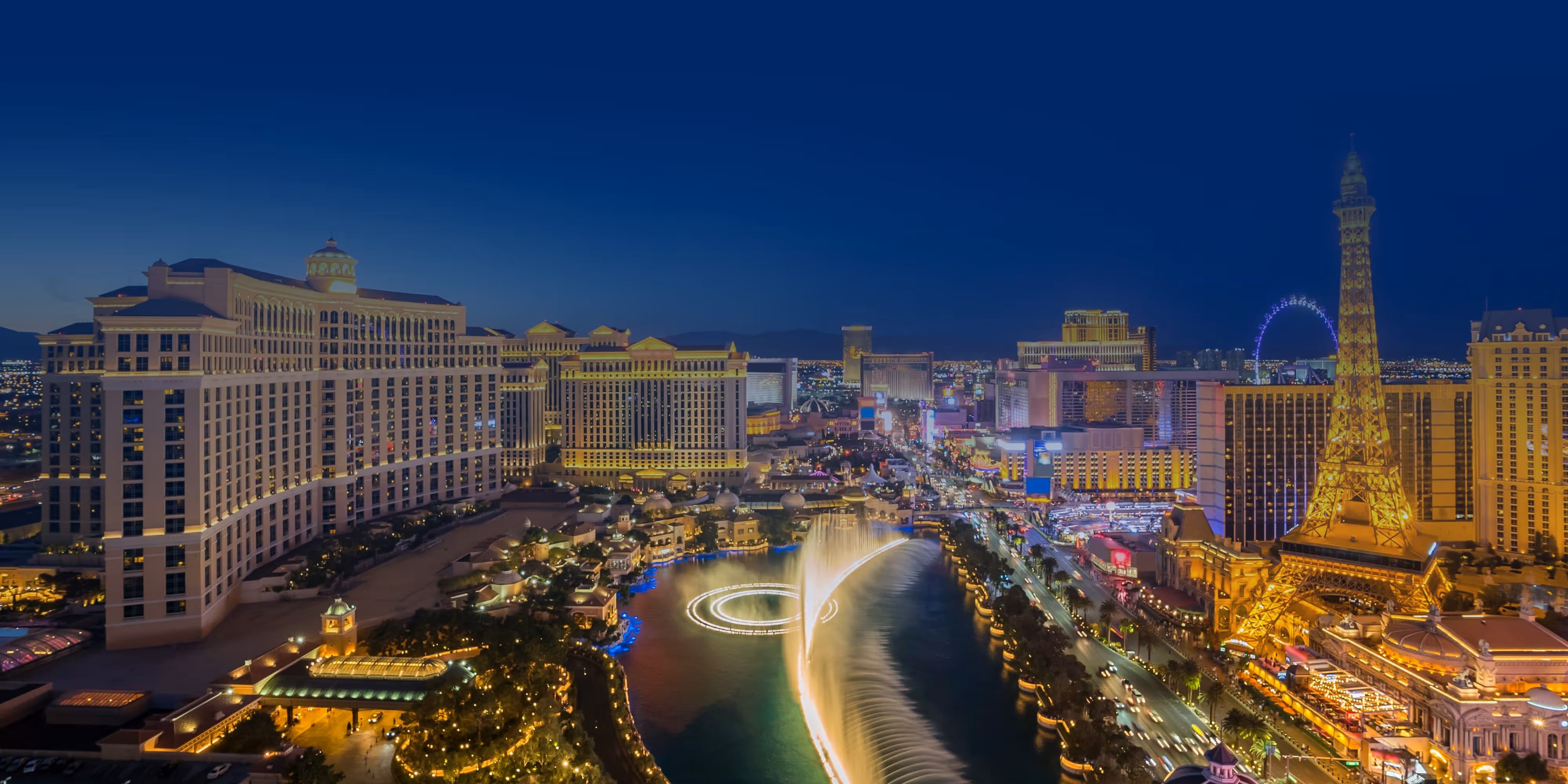 Aerial night view of Las Vegas Strip with Bellagio fountains, Eiffel Tower replica, and city lights.