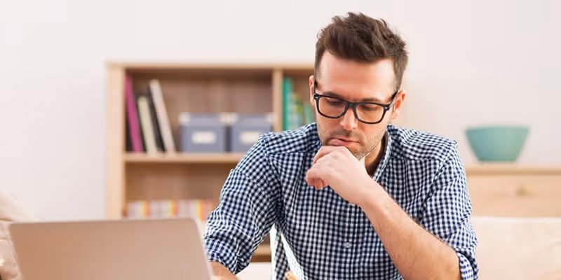 Man in glasses and checkered shirt sitting at desk, thinking while looking at laptop.