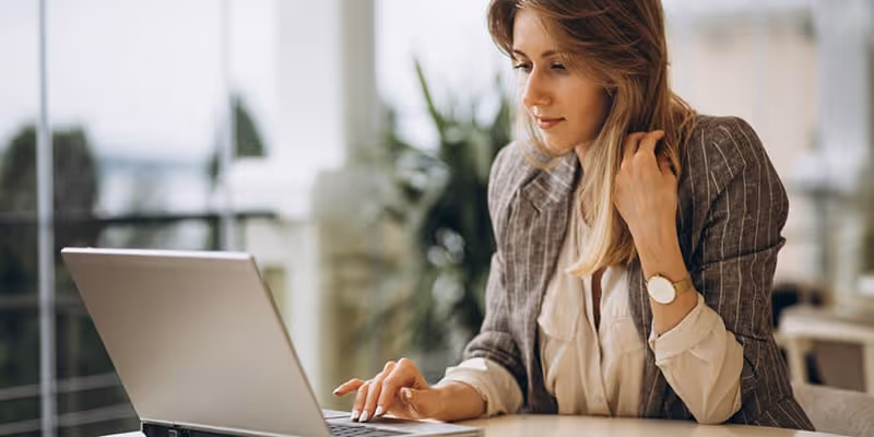 Woman wearing blazer working on her laptop at a desk in a bright office space.