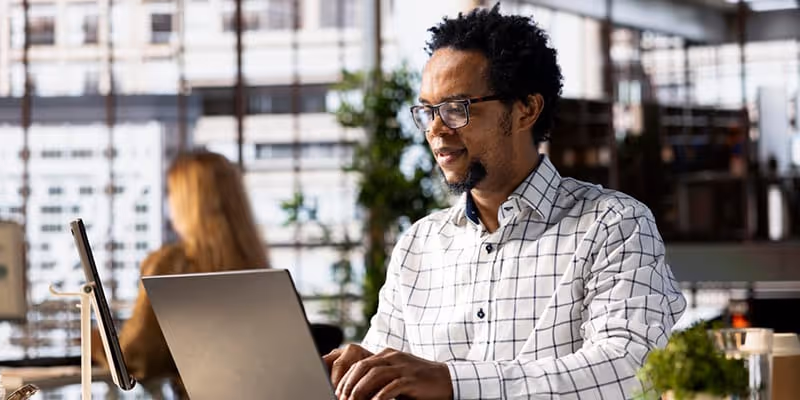 Man in glasses and checkered shirt typing on laptop in a bright office with plants and windows.