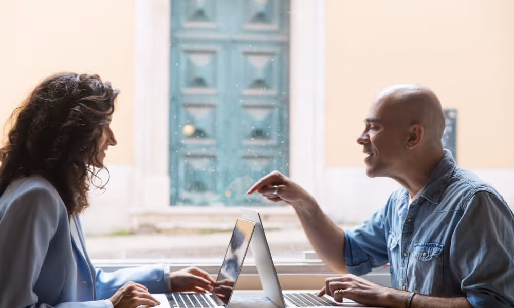 Two professionals discuss business strategy while working on laptops at a table.