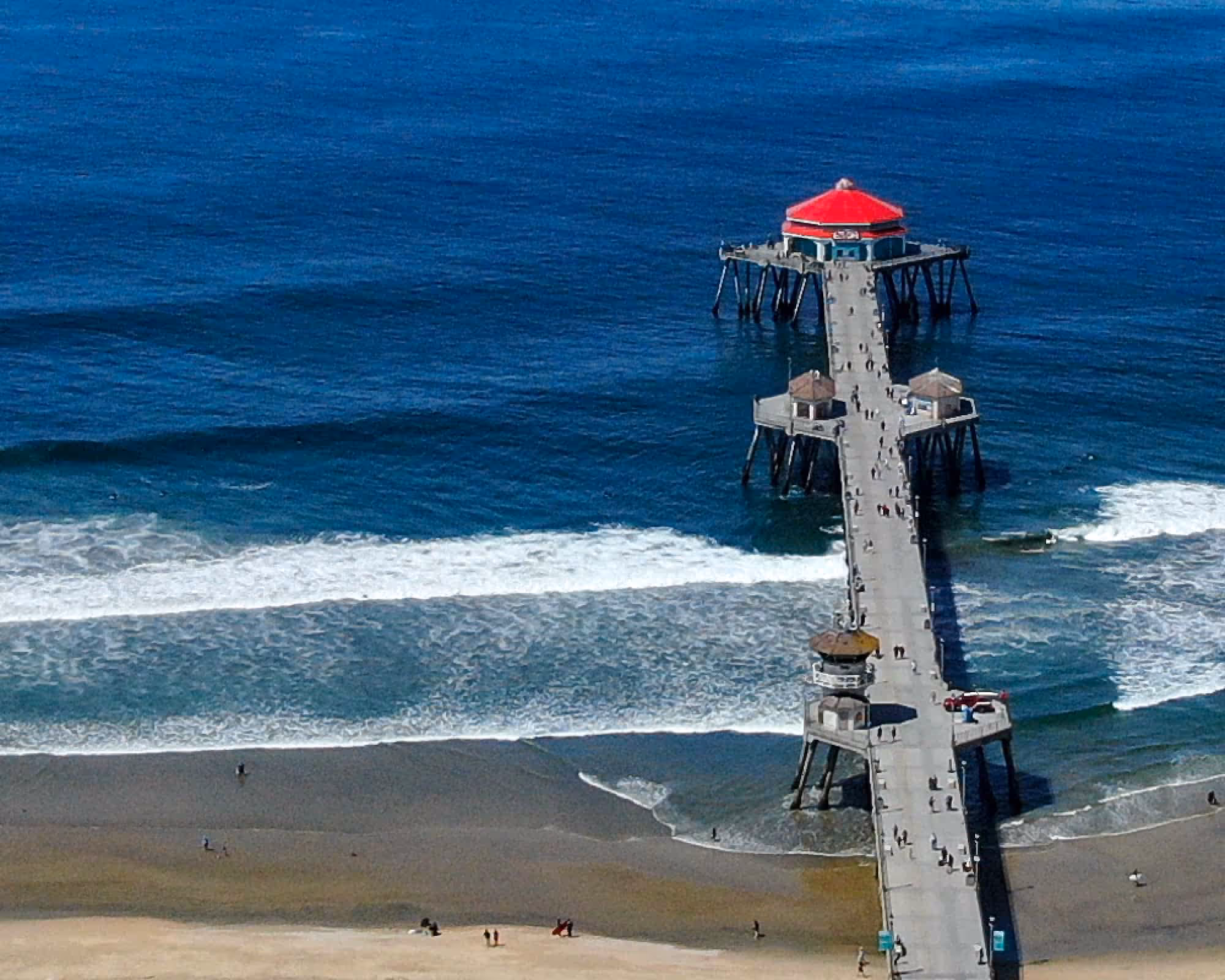A long pier with a red-roofed building extends over the ocean waves onto sandy beach.
