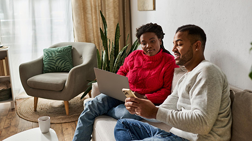 A couple sitting on a couch looking at a laptop and a mobile phone.