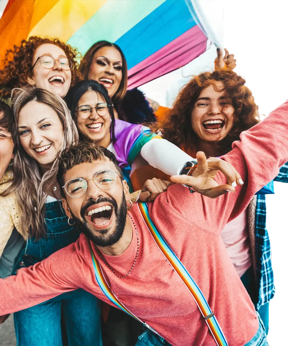 A diverse group of seven cheerful people huddled closely, smiling and laughing under a large rainbow flag, exuding unity and joy.