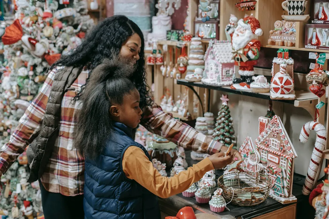 Free Young smiling ethnic woman with girl choosing gifts near Christmas tree in souvenir shop Stock Photo