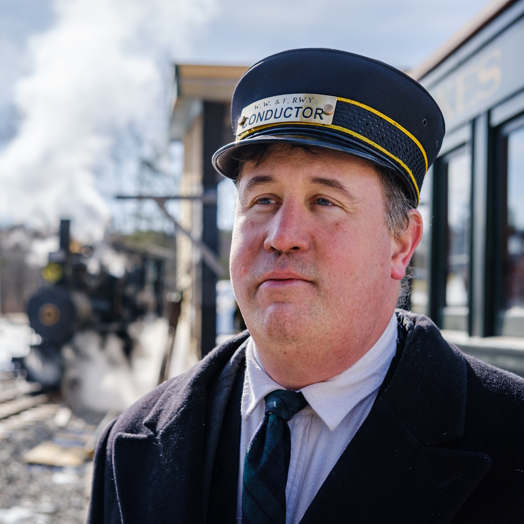 Ed Lecuyer in a WW&F Railway conductor's cap, with a steam locomotive behind him.