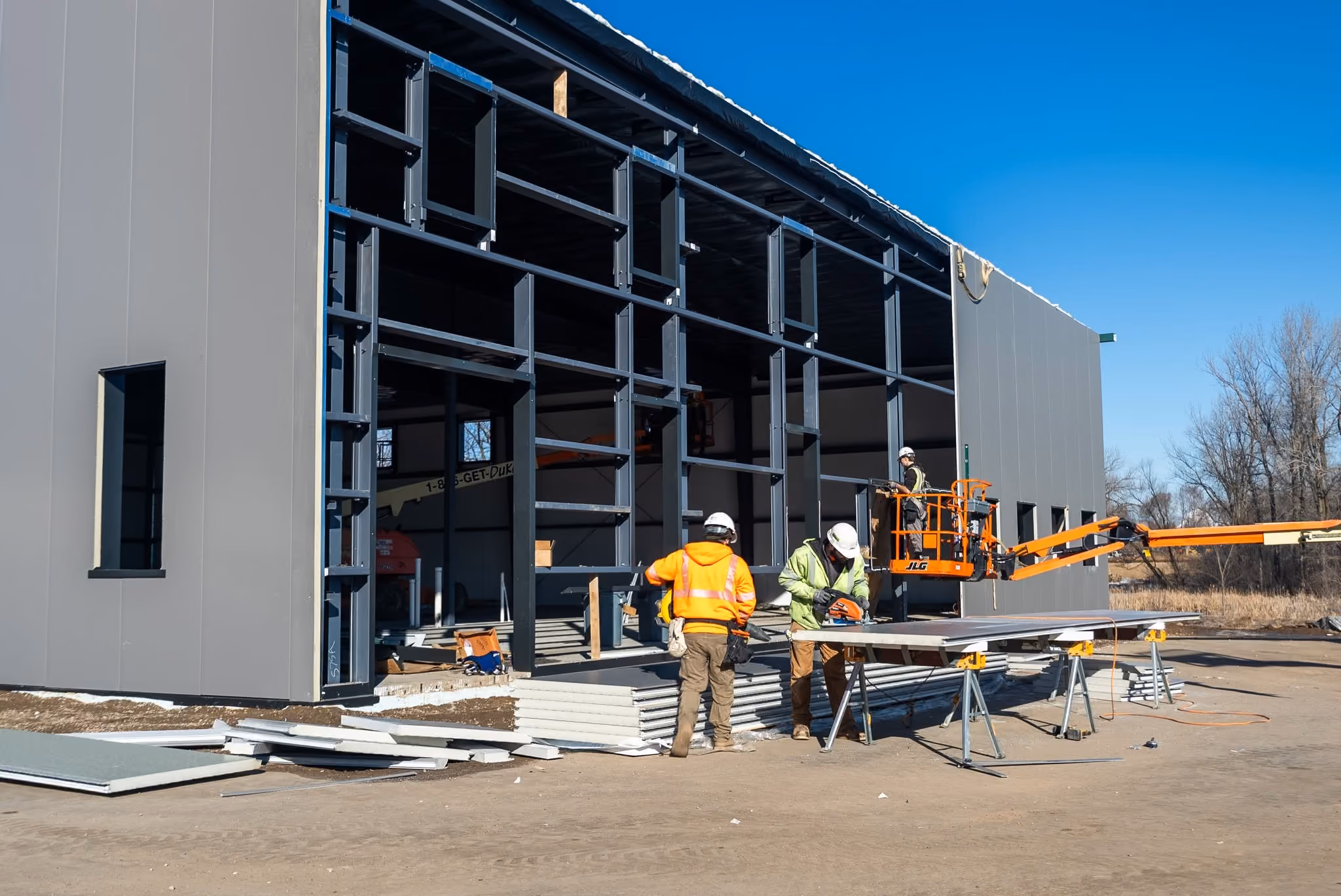 Country Creek Builders new Lakeville MN showroom under construction, workers installing exterior wall panels on steel framing with a JLG lift on site during winter framing phase