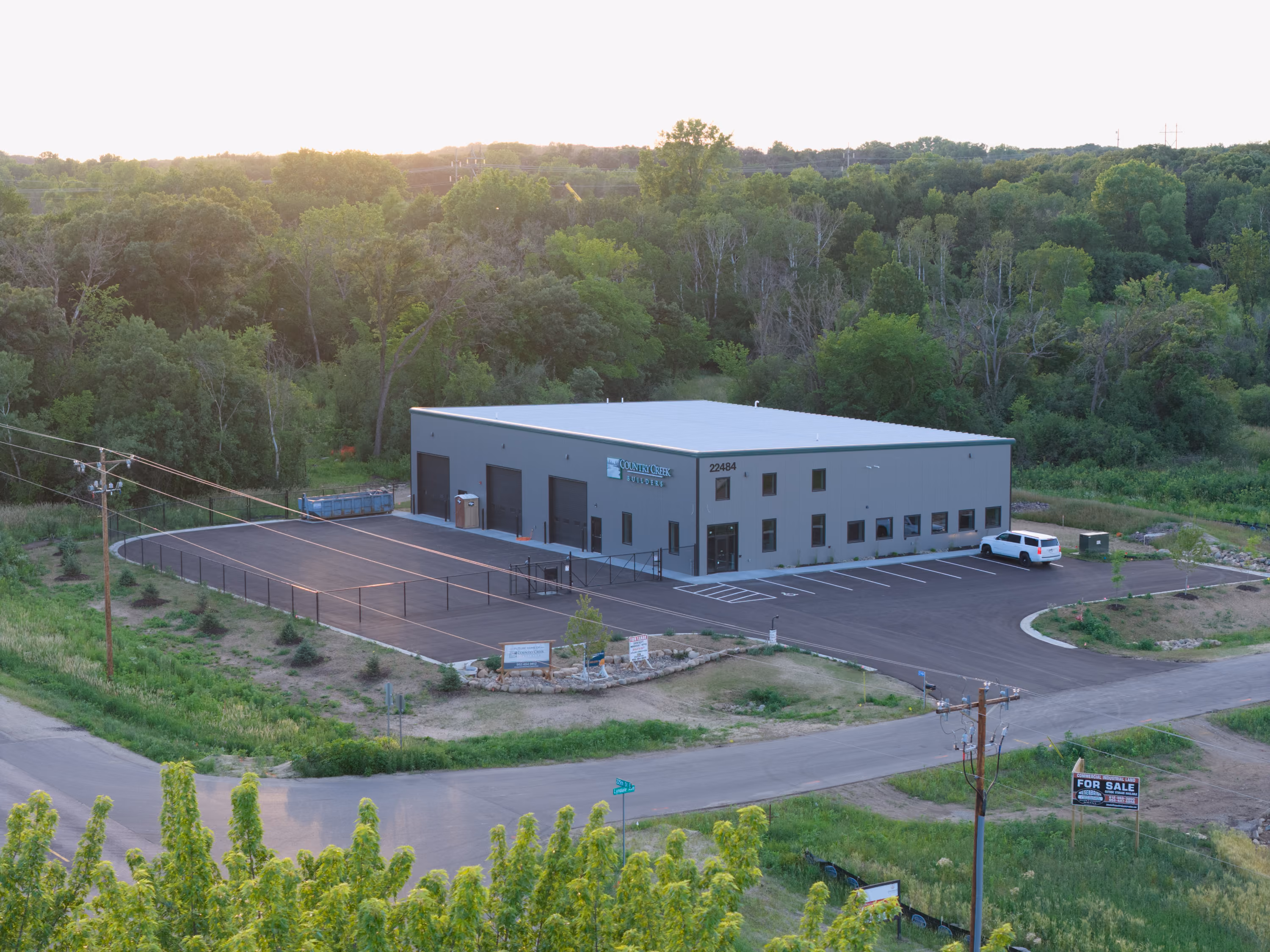 Country Creek Builders new Lakeville MN showroom building exterior, aerial view at dusk showing completed gray metal panel construction with green trim, parking lot, and wooded surroundings